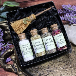 May include: A black gift box containing four small glass jars with cork stoppers and a miniature straw broom with a gold crescent moon charm. The jars are filled with white salt, dried rosemary, dried lavender, and dried rose petals.