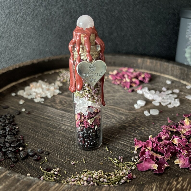 May include: A small glass bottle filled with dried herbs, rose petals, and crystals. The bottle is sealed with red wax and a heart-shaped charm that reads "Blessed Be".