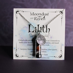 K&ouml;nnte beinhalten: Eine silberne Halskette mit einem Glas-Anh&auml;nger, der verschiedene Kristalle und einen silbernen Anh&auml;nger mit einem Symbol enth&auml;lt. Die Halskette wird auf einer wei&szlig;en Karte mit dem Text "Moondust and Raven Lilith Spellulet" und einer Beschreibung der Halskette pr&auml;sentiert.