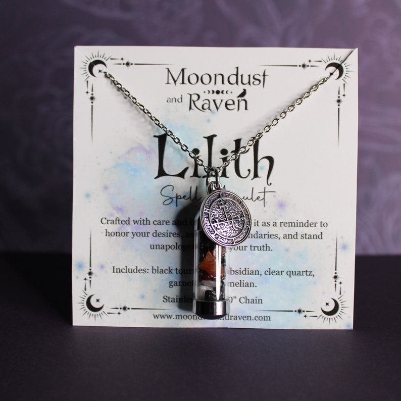 K&ouml;nnte beinhalten: Eine silberne Halskette mit einem Glas-Anh&auml;nger, der verschiedene Kristalle und einen silbernen Anh&auml;nger mit einem Symbol enth&auml;lt. Die Halskette wird auf einer wei&szlig;en Karte mit dem Text "Moondust and Raven Lilith Spellulet" und einer Beschreibung der Halskette pr&auml;sentiert.