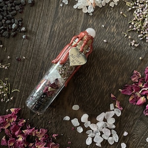 May include: A glass bottle filled with dried herbs, rose petals, and crystals. The bottle is sealed with a cork and has a red wax seal. A silver heart charm with the words "Blessed Be" hangs from the bottle.