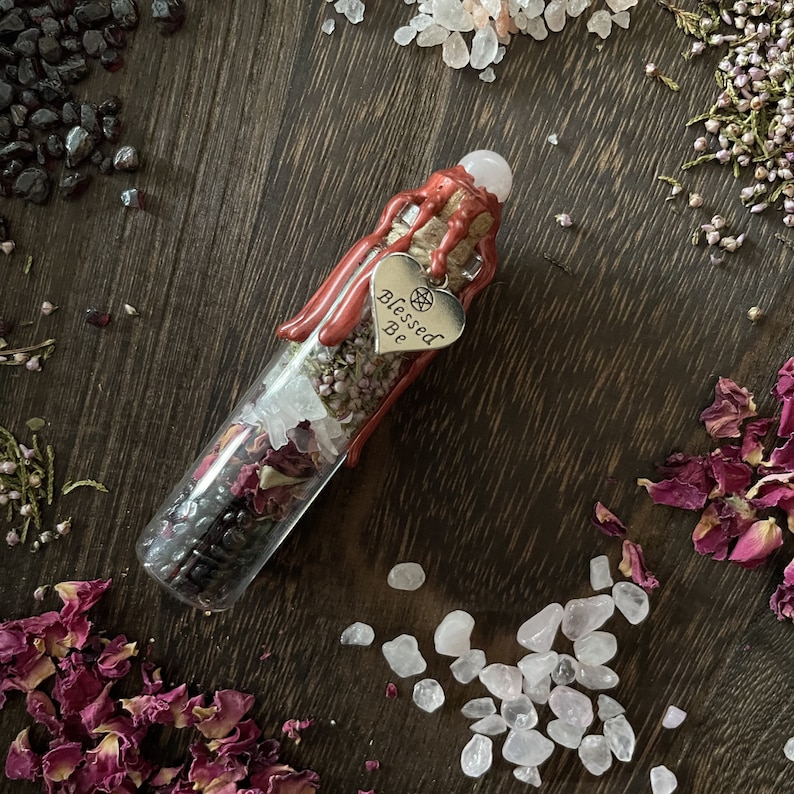 May include: A glass bottle filled with dried herbs, rose petals, and crystals. The bottle is sealed with a cork and has a red wax seal. A silver heart charm with the words "Blessed Be" hangs from the bottle.