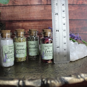 May include: Four small glass jars with cork stoppers, each containing dried herbs or salt and a label with text. A metal ruler placed next to the jars shows they are approximately 2.5 inches tall. The labels feature phrases like Always Throw Spilled Salt, Keep Rosemary, Plant Lavender, and And Fall In Love.