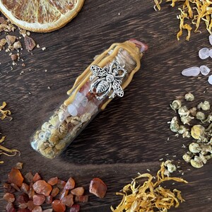 May include: A glass bottle filled with dried flowers, crystals, and honey. The bottle is topped with a silver bee charm and a wax seal. The bottle is surrounded by dried flowers and crystals.