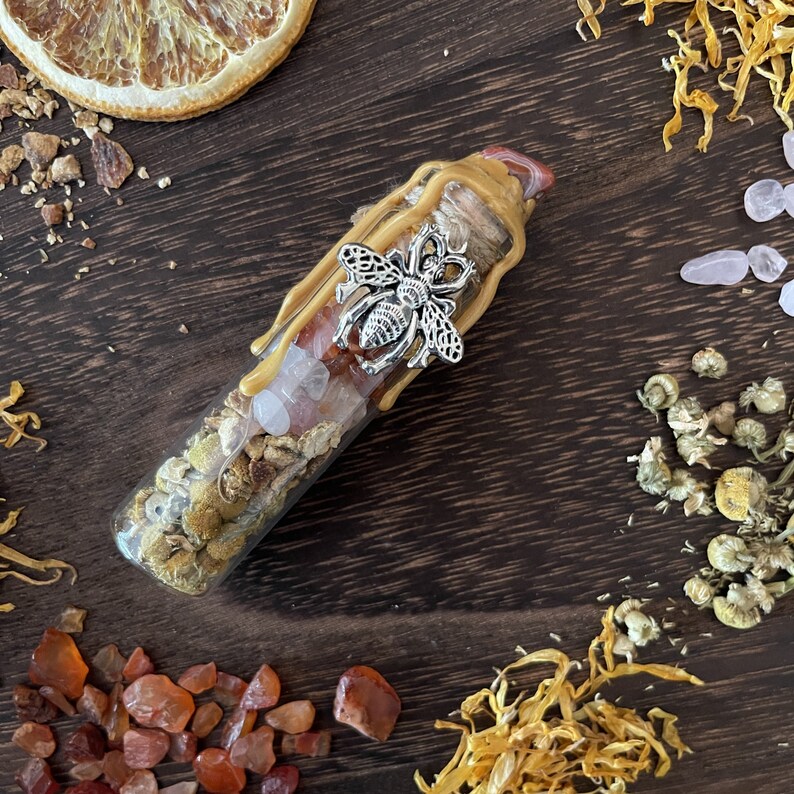 May include: A glass bottle filled with dried flowers, crystals, and honey. The bottle is topped with a silver bee charm and a wax seal. The bottle is surrounded by dried flowers and crystals.