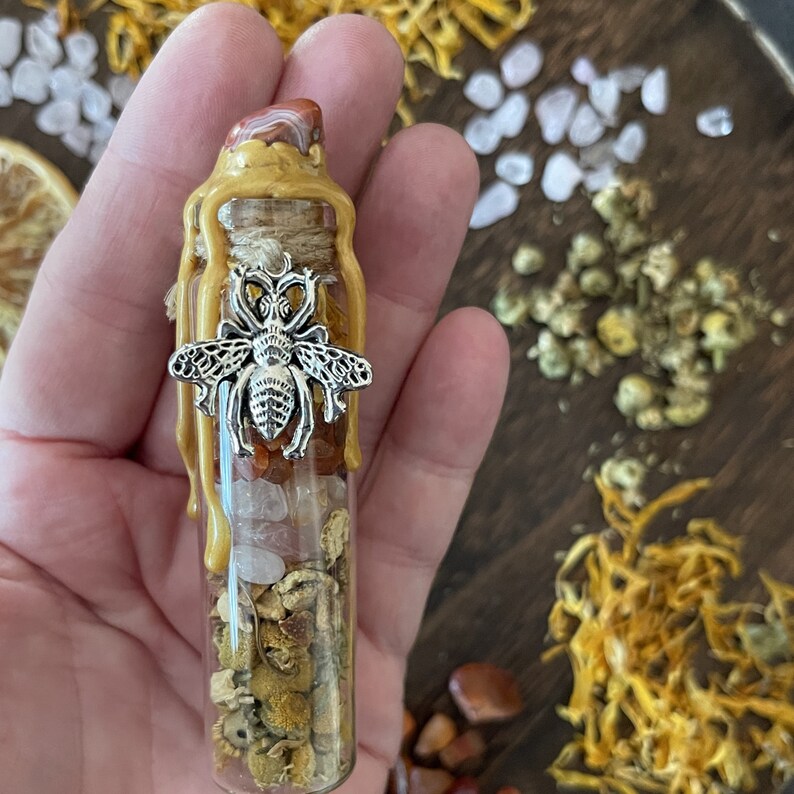 May include: A small glass bottle filled with various crystals, herbs, and a bee charm. The bottle is sealed with gold wax and has a red and white striped stopper.