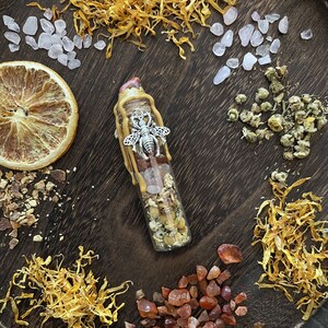 May include: A small glass bottle filled with dried flowers, crystals, and herbs. The bottle is topped with a wax seal and a silver bee charm. The bottle is surrounded by dried flowers, crystals, and herbs.