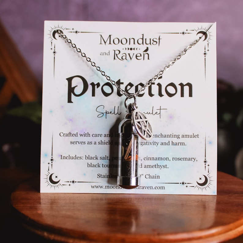 Puede incluir: Un collar de plata con un colgante de vial de vidrio lleno de hierbas y cristales. El colgante tiene un amuleto de pentagrama de plata. La tarjeta dice "Moondust and Raven Protection Spell Amulet".