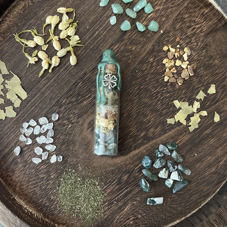 K&ouml;nnte beinhalten: Eine gr&uuml;ne Glasflasche gef&uuml;llt mit getrockneten Kr&auml;utern, Kristallen und einem Gl&uuml;cksbringer mit vierbl&auml;ttrigem Kleeblatt. Die Flasche ist umgeben von verschiedenen getrockneten Kr&auml;utern, Kristallen und anderen nat&uuml;rlichen Elementen.