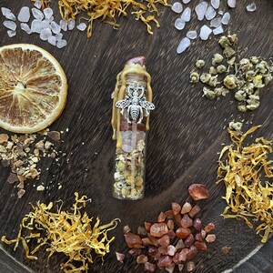 May include: A clear glass bottle filled with crystals, dried flowers, and a silver bee charm. The bottle is topped with a gold wax seal and sits on a wooden surface with dried flowers and crystals.