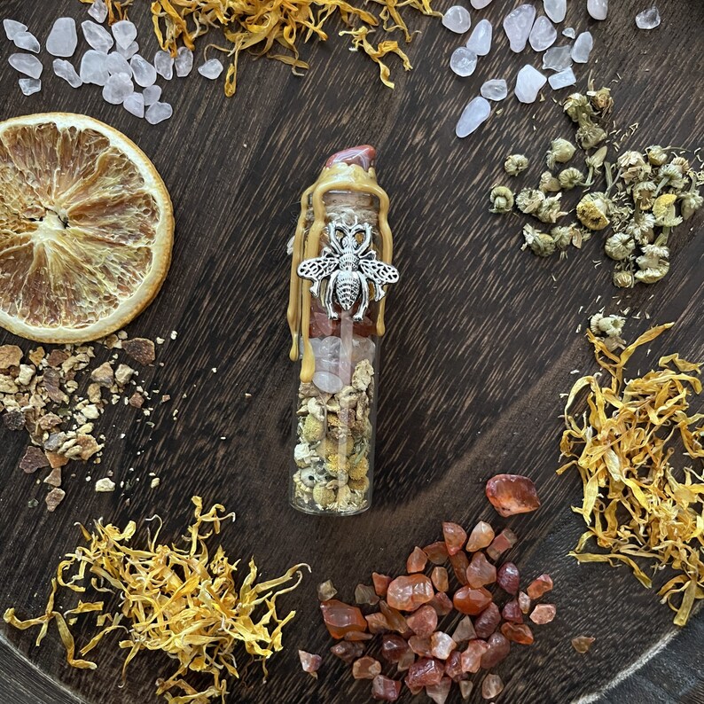 May include: A clear glass bottle filled with crystals, dried flowers, and a silver bee charm. The bottle is topped with a gold wax seal and sits on a wooden surface with dried flowers and crystals.