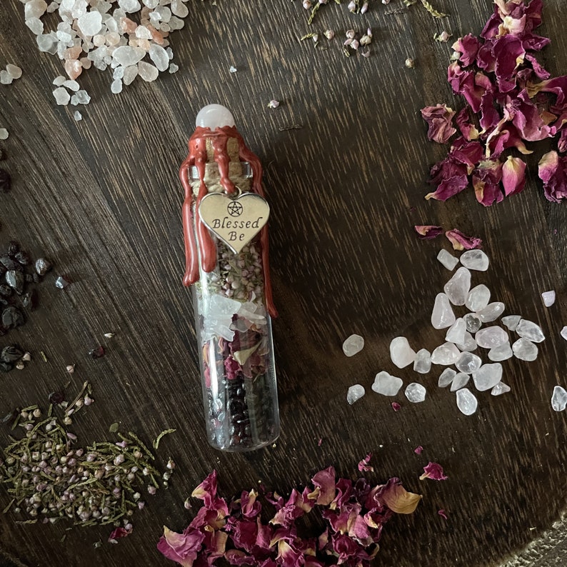 May include: A small glass bottle filled with dried herbs, crystals, and rose petals. The bottle is sealed with red wax and has a silver charm that says "Blessed Be".