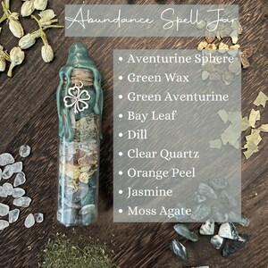 K&ouml;nnte beinhalten: Ein gr&uuml;nes Glasgef&auml;&szlig; gef&uuml;llt mit Kristallen, Kr&auml;utern und einem gr&uuml;nen Wachssiegel. Das Gef&auml;&szlig; ist mit "Abundance Spell Jar" beschriftet und listet die Zutaten auf: Aventurin-Kugel, gr&uuml;nes Wachs, gr&uuml;ner Aventurin, Lorbeerblatt, Dill, klarer Quarz, Orangenschale, Jasmin und Moosachat.