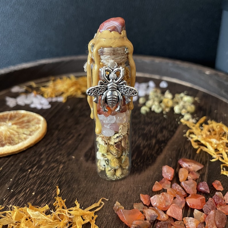 May include: A small glass bottle filled with various crystals, dried flowers, and herbs. The bottle is topped with a wax seal and a silver bee charm. The bottle is sitting on a wooden surface with orange slices and other natural elements.