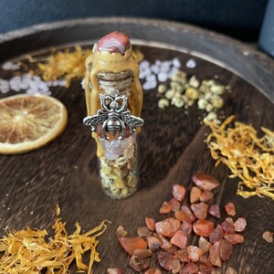 May include: A small glass bottle filled with crystals, dried flowers, and herbs. The bottle is topped with a wax seal and a bee charm. The bottle is surrounded by orange and red crystals.