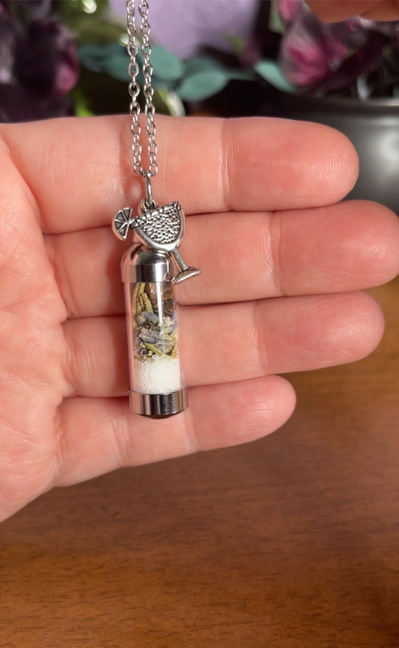 Puede incluir: Collar de plata con un vial de vidrio lleno de hierbas secas y un dije de plata de un vaso de margarita.