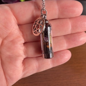Puede incluir: Un collar de cadena plateada con un colgante de vial de vidrio transparente lleno de cristales y hierbas. El vial est&aacute; coronado con una tapa plateada y un amuleto de pentagrama plateado cuelga de la tapa.