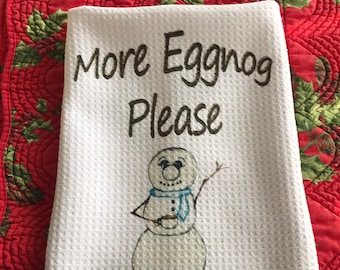 以下が含まれることがあります： 雪だるまがマグカップを持っている白いキッチンクロスで、「More Eggnog Please」という文字が刺繍されています。