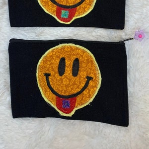 May include: Two black zippered pouches with a yellow smiley face with a red tongue and a blue and red pill on it. The pouches have a floral pattern inside the smiley face.