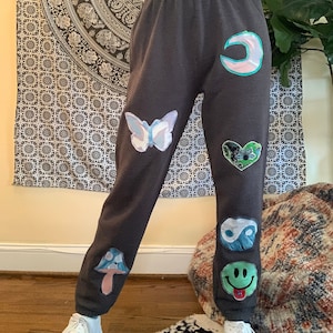 Puede incluir: Pantalones de chándal grises con varios parches coloridos cosidos, incluyendo una mariposa, una luna creciente, un corazón, un símbolo yin yang, un hongo y una cara sonriente.