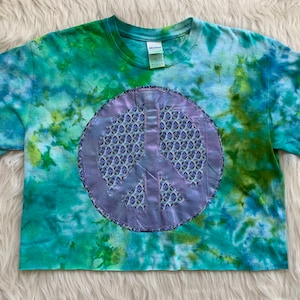 May include: A tie-dye cropped t-shirt featuring a purple peace sign with a blue and white geometric pattern. The shirt is a light blue, green, and white colour.