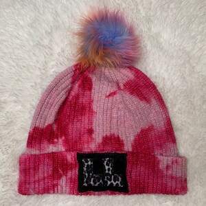 Pode incluir: Boina tie-dye rosa e roxo com um remendo preto com uma letra "L" branca e um pompom preto no topo.