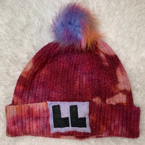 May include: A red, pink, and orange tie-dye beanie with a purple and blue pom pom on top. The beanie has a black patch with the letters "LL" in white on the front.