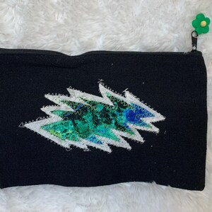 May include: Black canvas zippered pouch with a green, blue, and white lightning bolt design. The zipper pull is a green flower.