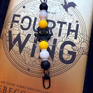 May include: A beaded lanyard with a black clasp, featuring black, white, and yellow beads. A black dragon charm is centered on the lanyard. The lanyard is placed on a book with the title "Fourth Wing" and the author's name, Rebecca Yarros.