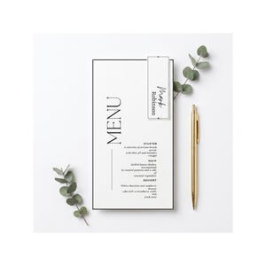 May include: A rectangular white menu card with black text featuring the word MENU, a starter, main, and dessert section, and a name tag reading Mark Robinson. A gold pen and two sprigs of green eucalyptus leaves are placed beside the card.