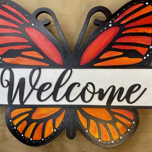 May include: A wooden welcome sign shaped like a butterfly with orange and black wings. The sign has the word "Welcome" written in black cursive lettering on a white background.