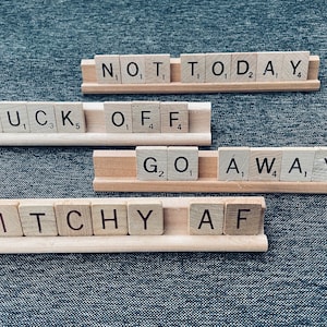 以下が含まれることがあります： 「NOT TODAY」、「FUCK OFF」、「GO AWAY」、および「BITCHY AF」などのフレーズを綴る木製の文字タイルが、木製のタイルホルダーに配置されています。 タイルは、テクスチャ加工された濃い青色の表面にあります。