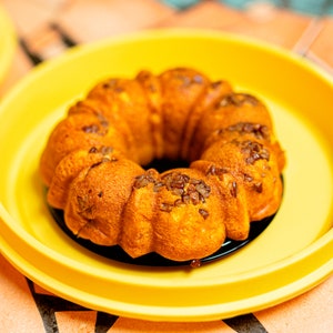 以下が含まれることがあります： 輪の形をした、金茶色のバントケーキ。刻んだナッツがトッピングされ、黒いお皿にのせられ、明るい黄色の皿の中に置かれています。ケーキは質感のある表面をしています。