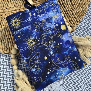 Könnte beinhalten: Eine blaue Stofftasche mit einem goldenen Konstellationsdesign mit Sternen, Monden und Tieren. Die Tasche hat einen schwarzen Kordelverschluss und einen goldenen Quasten.