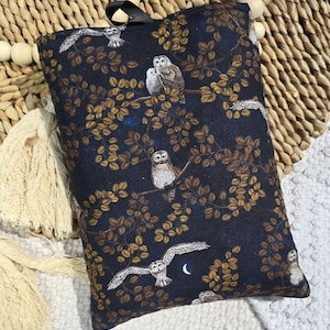 May include: A dark blue book sleeve with a repeating pattern of owls on branches with golden leaves. The owls are in white, brown, and grey. A black ribbon is attached to the top for carrying.