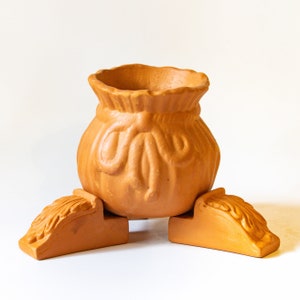 May include: A terracotta planter shaped like a bag with a bow, standing on three legs. The planter is a light brown color and has a rustic, handmade look.