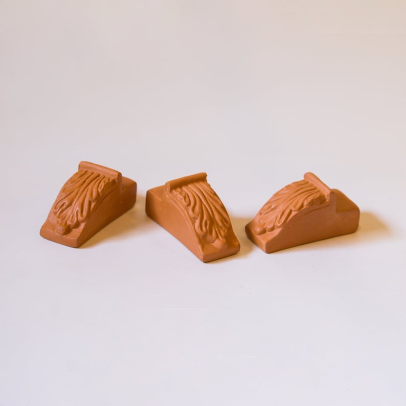 May include: Three terracotta-colored decorative corner brackets with an ornate design. The brackets are made of a textured material and have a slightly curved shape.