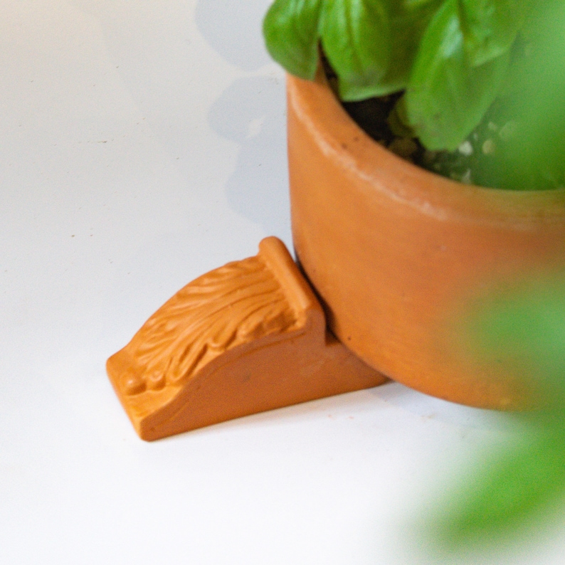 May include: A terracotta planter with a green plant inside, sitting on a terracotta plant stand with an ornate design.