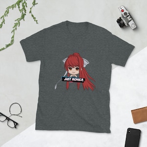 Puede incluir: Camiseta gris oscuro jaspeada con una imagen de dibujos animados de una niña con cabello rojo y un lazo blanco. El texto "JUST MONICA" está impreso en una fuente colorida y estilizada sobre una barra negra.
