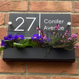 May include: A black and white house number sign with the number 27 and the street name Conifer Avenue. The sign is mounted on a brick wall with a black planter box underneath filled with purple and pink flowers.