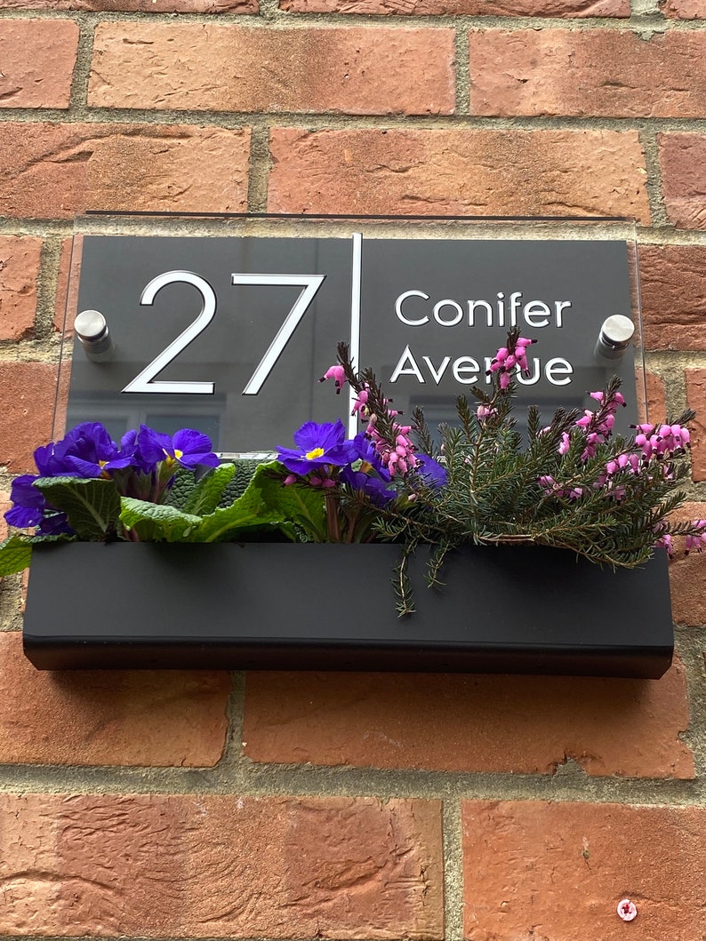 May include: A black and white house number sign with the number 27 and the street name Conifer Avenue. The sign is mounted on a brick wall with a black planter box underneath filled with purple and pink flowers.