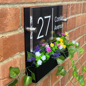 May include: Black acrylic house number sign with white numbers and text. The sign reads "27 Conifer Avenue". The sign is mounted on a brick wall with a black planter box underneath. The planter box is filled with artificial flowers.
