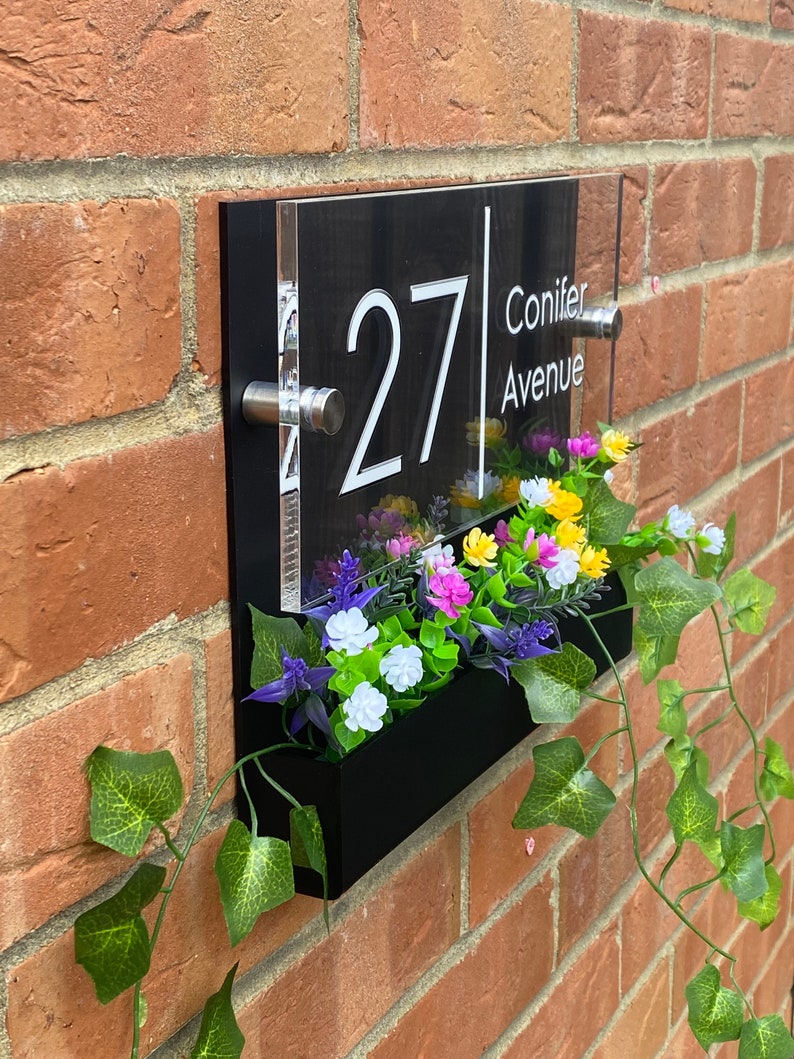 May include: Black acrylic house number sign with white numbers and text. The sign reads "27 Conifer Avenue". The sign is mounted on a brick wall with a black planter box underneath. The planter box is filled with artificial flowers.