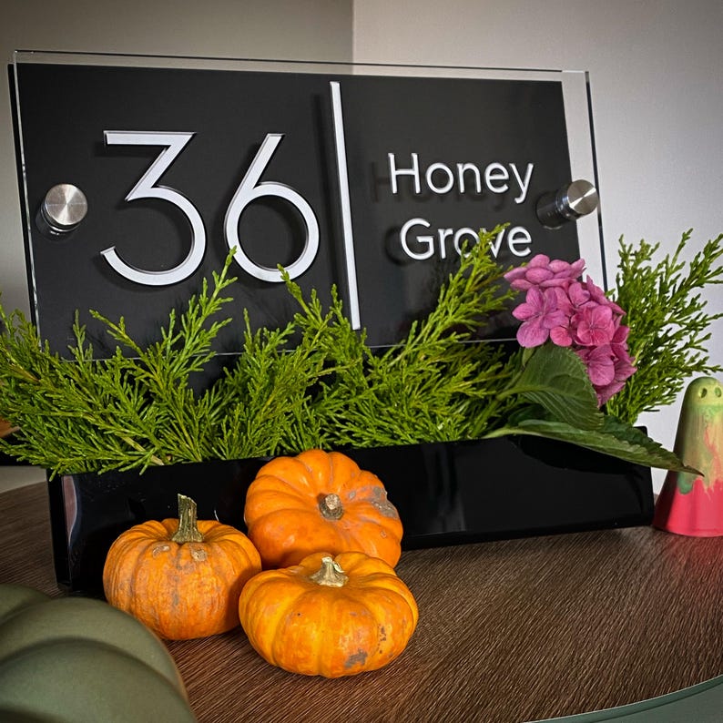 May include: Black acrylic house number sign with white numbers "36" and the text "Honey Grove". The sign is mounted on a black stand with a potted plant in front of it. Three small orange pumpkins are in the foreground.