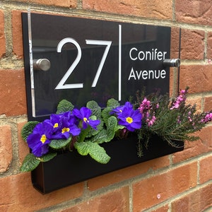 May include: Black acrylic house number sign with white lettering. The sign reads "27 Conifer Avenue". The sign is mounted on a brick wall with a black planter box underneath.