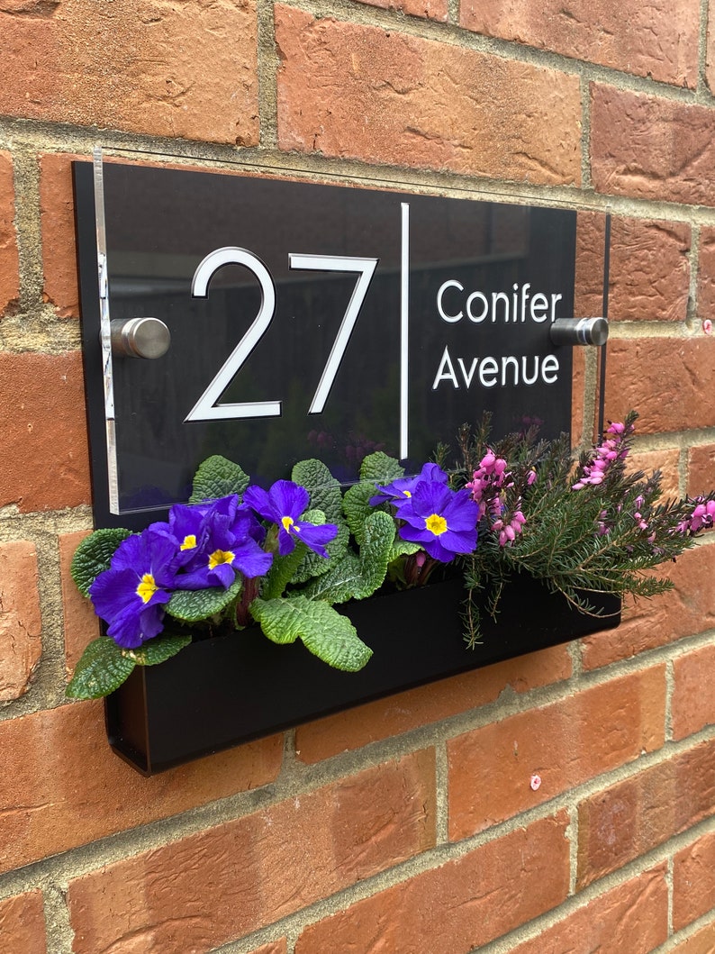 May include: Black acrylic house number sign with white lettering. The sign reads "27 Conifer Avenue". The sign is mounted on a brick wall with a black planter box underneath.
