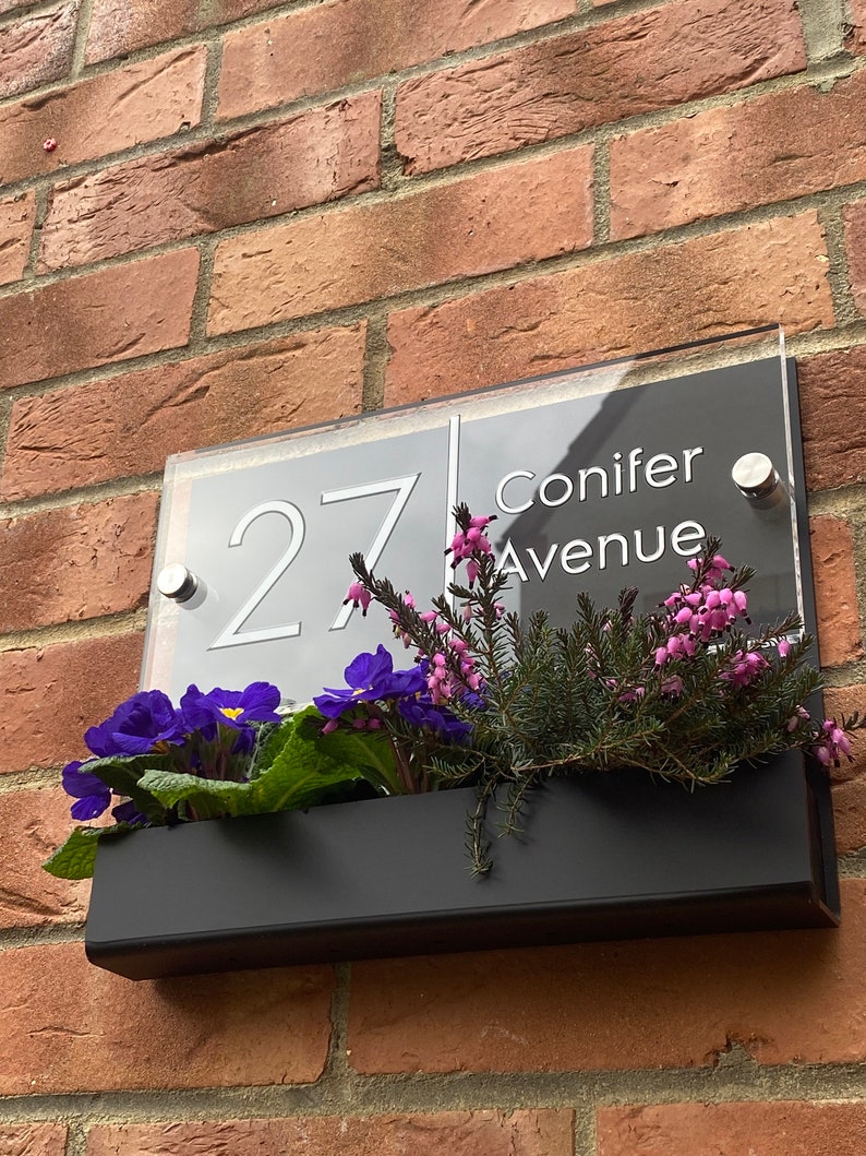 May include: A black and white house number sign with the number 27 and the street name Conifer Avenue. The sign is mounted on a brick wall with a black planter box underneath filled with pink and purple flowers.