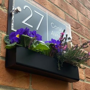 May include: A black acrylic house number sign with white numbers and text. The sign reads "27 Conifer Ave". The sign is mounted on a brick wall with a black planter box underneath.