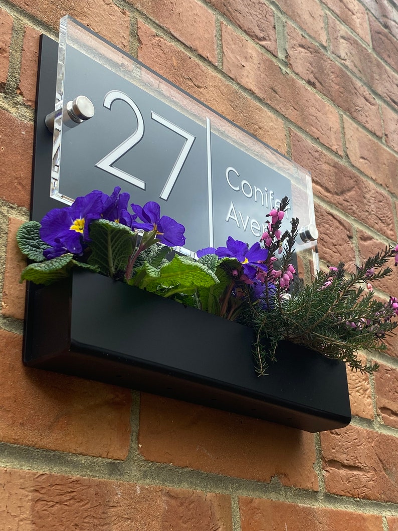 May include: A black acrylic house number sign with white numbers and text. The sign reads "27 Conifer Ave". The sign is mounted on a brick wall with a black planter box underneath.