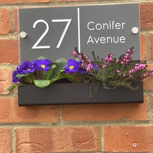 May include: A gray acrylic house number sign with white lettering. The sign reads "27 Conifer Avenue". The sign is mounted on a brick wall with a black planter box underneath.